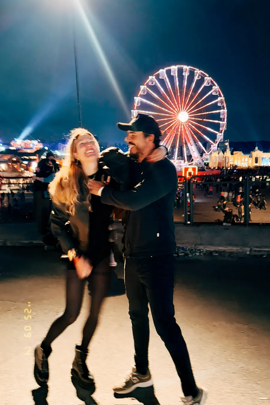 Gabriela Morais rindo abraçada ao companheiro em frente a uma roda-gigante iluminada.
